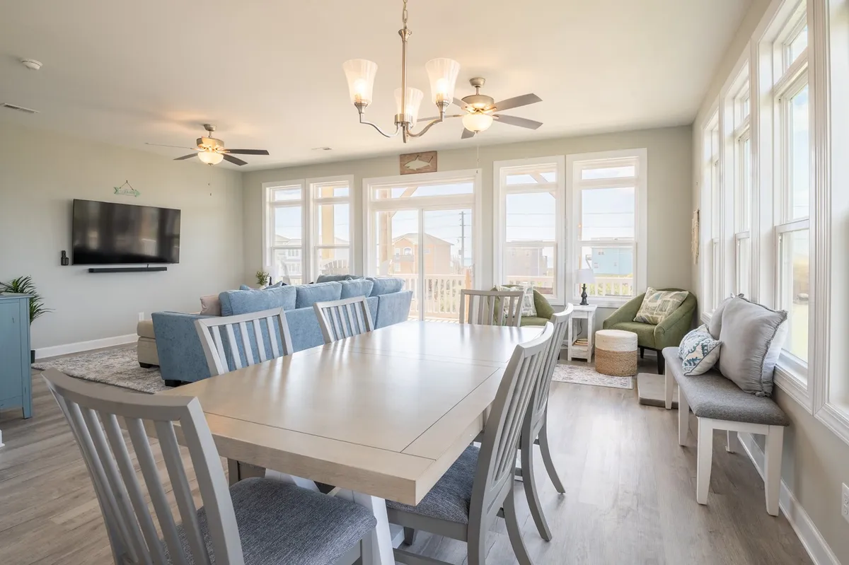 An open-concept living and dining area featuring a comfortable sofa, a dining table set for meals, large windows, and a television. The room is bathed in natural light, and the seating arrangement encourages both relaxation and interaction, with views to the outdoors.