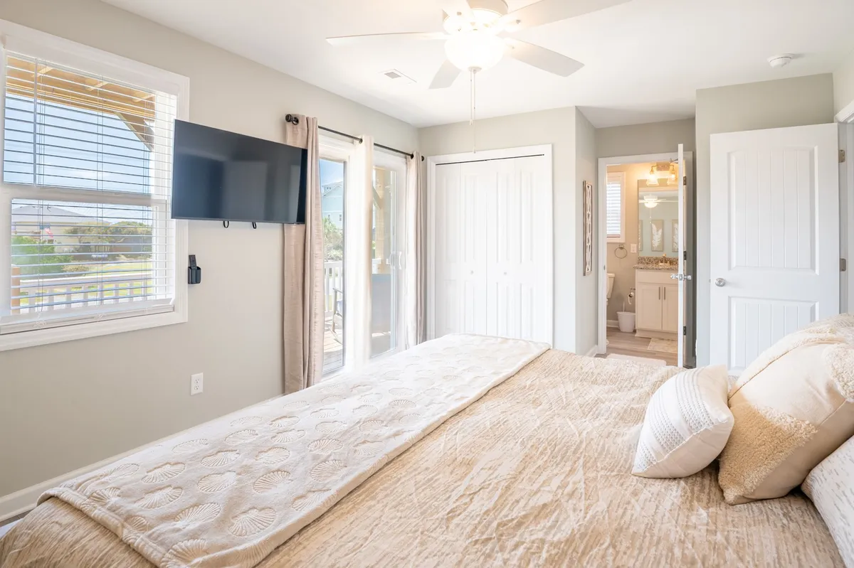 A cozy guest bedroom with cream-colored bedding on the bed, a wall-mounted TV, and sliding glass doors that open onto a private balcony. Natural light pours in, highlighting a comfortable space perfect for morning relaxation and enjoying the water view.