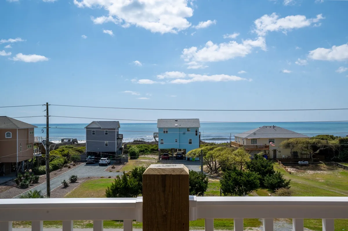 A serene view from a balcony showcasing the expansive Atlantic ocean, with neighboring houses nestled among greenery. The porch in the foreground invites relaxation while offering an unobstructed sunrise vista over the water.