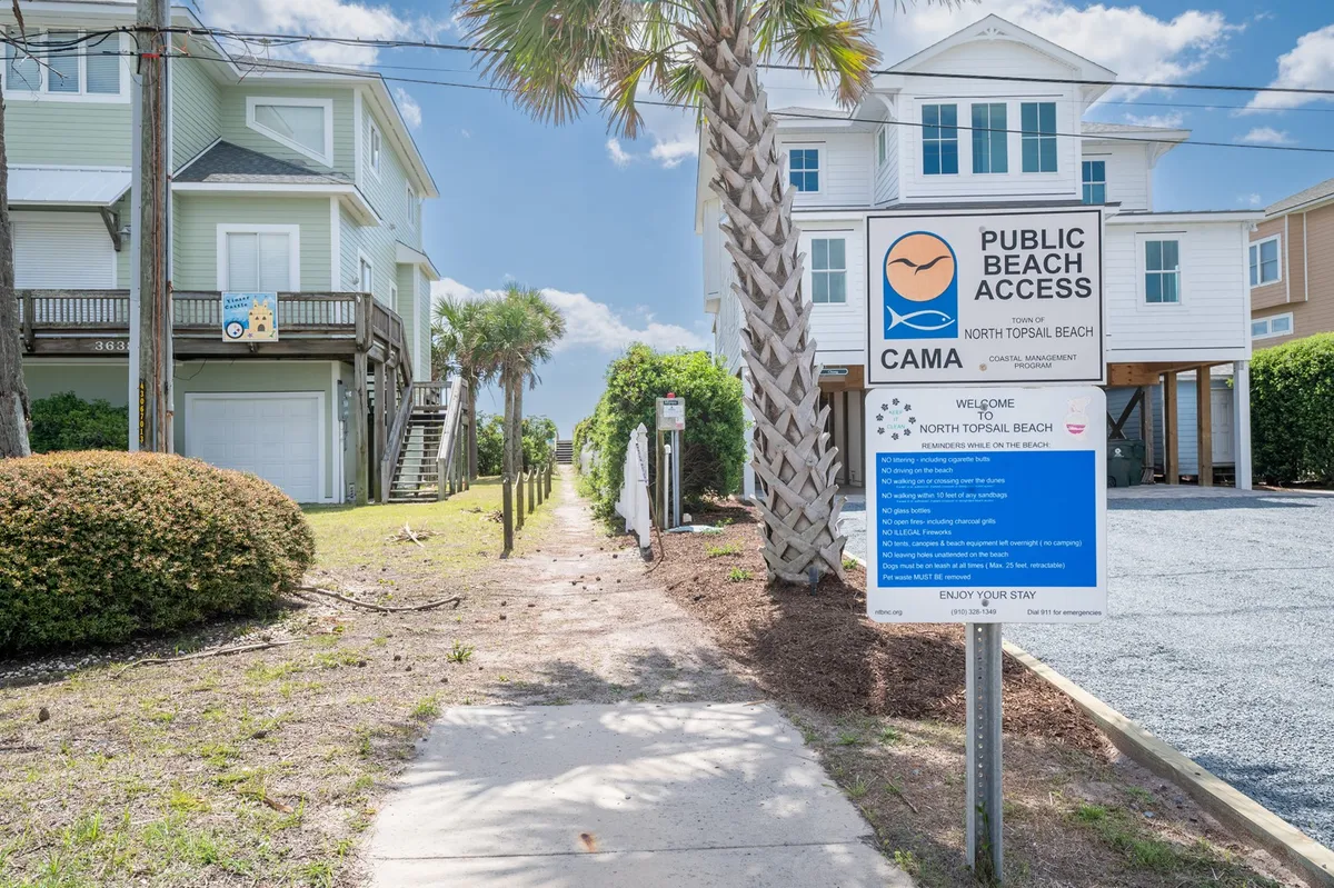A beach access sign stands at the front of a sandy pathway bordered by palm trees, leading through a coastal neighborhood towards the ocean. The sense of anticipation and ease is palpable, suggesting both accessibility and a welcoming community vibe.