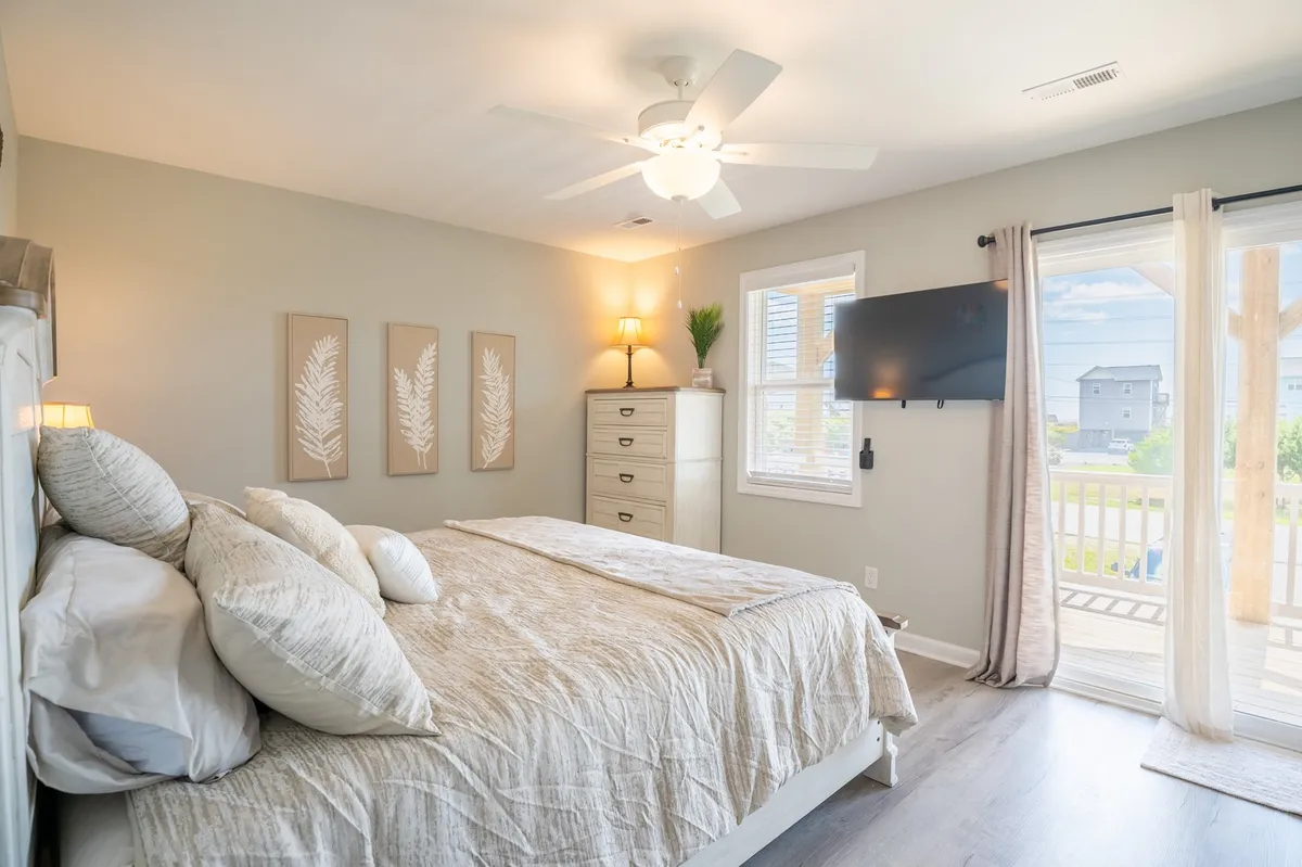 A relaxing bedroom featuring a bed positioned to take advantage of sunlight streaming in through the window, a ceiling fan overhead, a dresser, and wall art. The window offers views and access to an outdoor balcony, creating a tranquil atmosphere for guests.