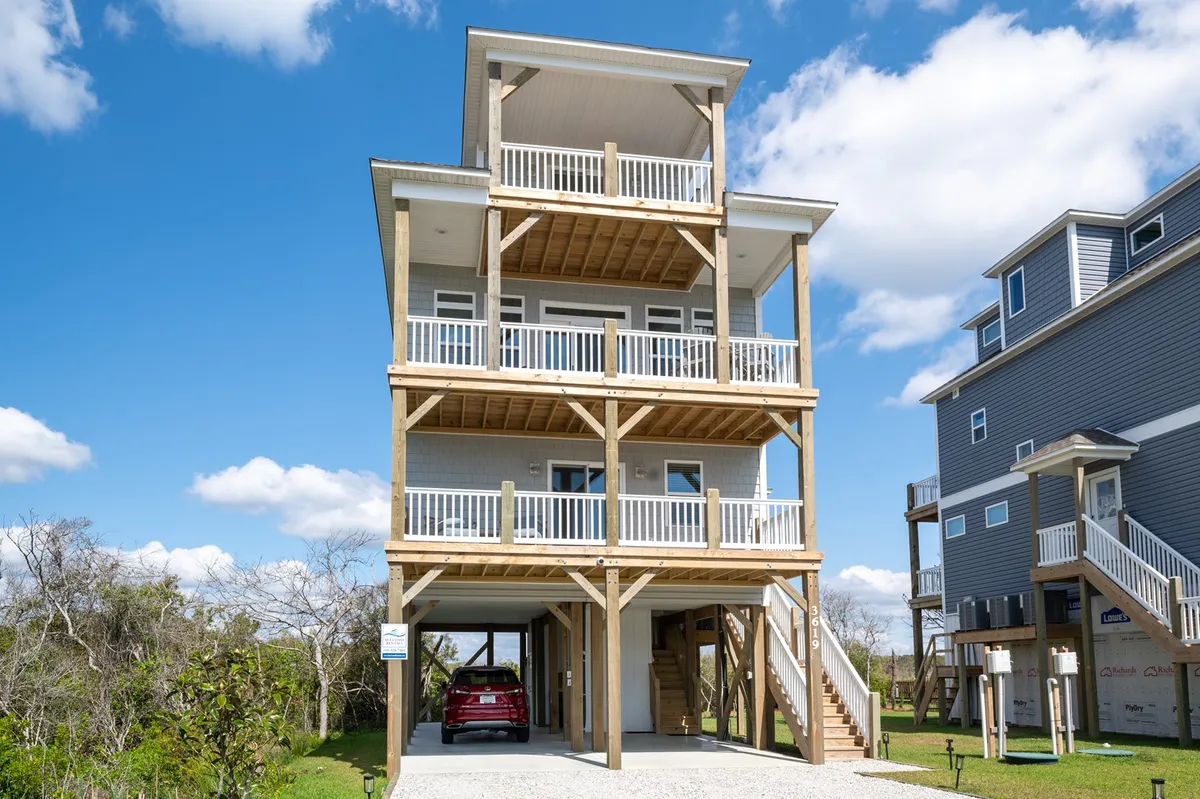 An exterior shot of a three-story beach house with multiple balconies, sunlit under a clear sky. The home’s architectural lines and outdoor spaces, including the upper balcony, clearly demonstrate the property’s elevated vantage points and outdoor living areas ideal for sunset viewing.