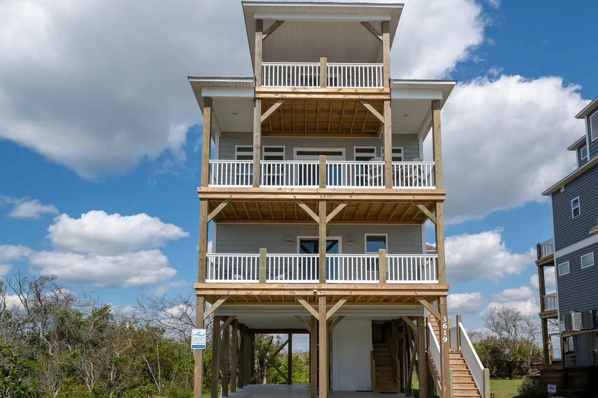The image showcases a stunning, brand-new three-story beach house in North Topsail Beach, with spacious balcony railings on each level, a staircase leading up to the main entrance, and inviting outdoor spaces perfect for relaxation. The view hints at the proximity to the water and promises gorgeous sunrises and breezy evenings.