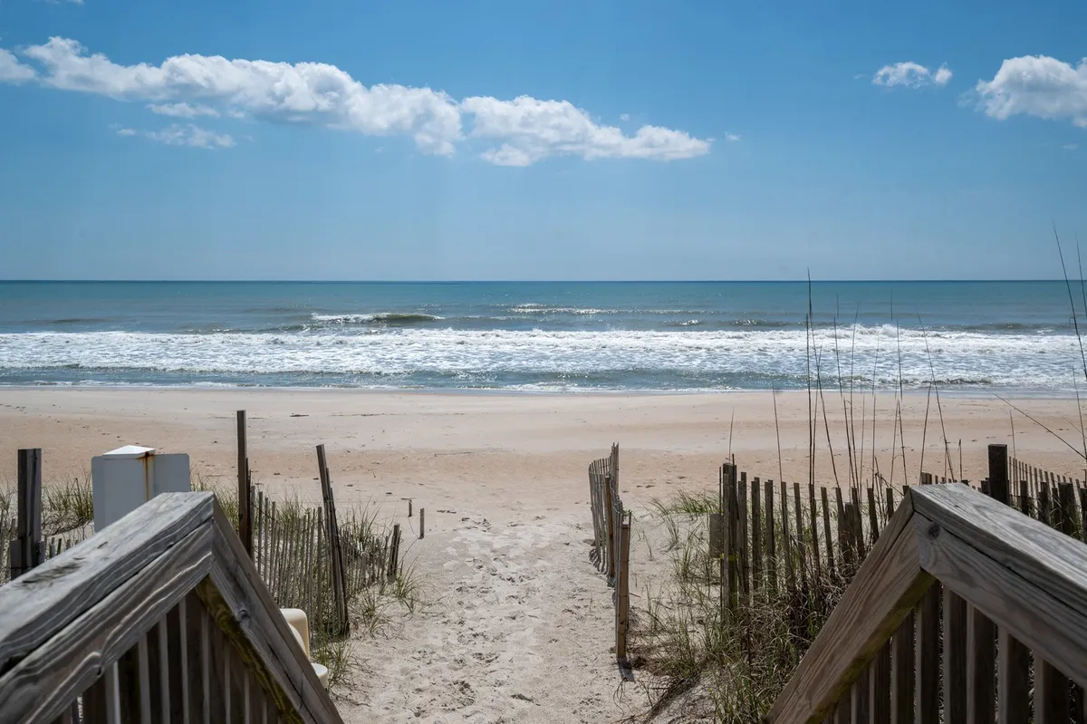 A wide, inviting sandy beach stretches out to meet gentle ocean waves, framed by dune grass and a wooden pathway leading directly onto the sand. The scene evokes the ideal family beach day, with open space for games, sunbathing, and waterside relaxation.