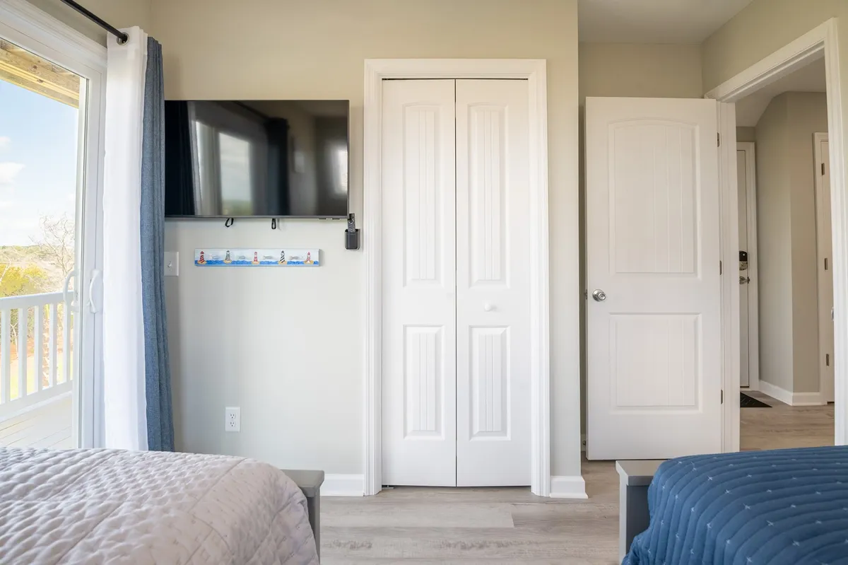 A spacious primary bedroom featuring a king bed, large windows that fill the space with natural light, a cozy sitting area with a TV, and closet doors visible. The windows offer serene views, creating a restful atmosphere ideal for a private retreat.