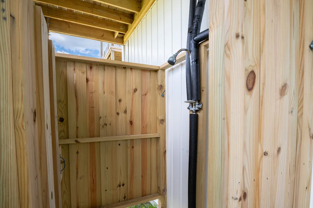 A refreshing outdoor shower is set against wooden fencing, open to the sky and surrounded by open space. The shower fixture is clearly visible, offering a practical post-beach rinse-off spot.