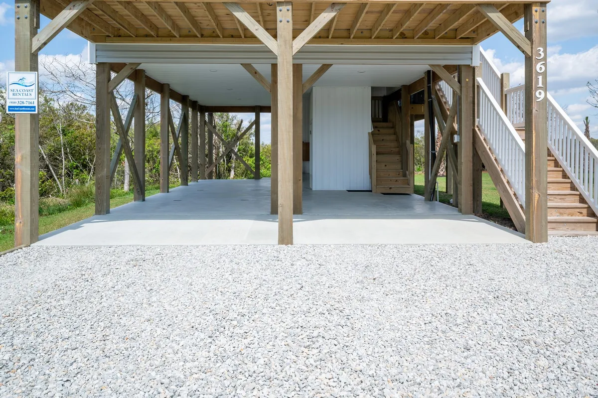 A relaxed exterior shot showing the gravel driveway and ample parking area beneath the house, supported by sturdy wooden beams. The staircase leads up to the main living area, and the shaded space under the house provides a cool spot ideal for prepping beach gear or letting a pet rest.